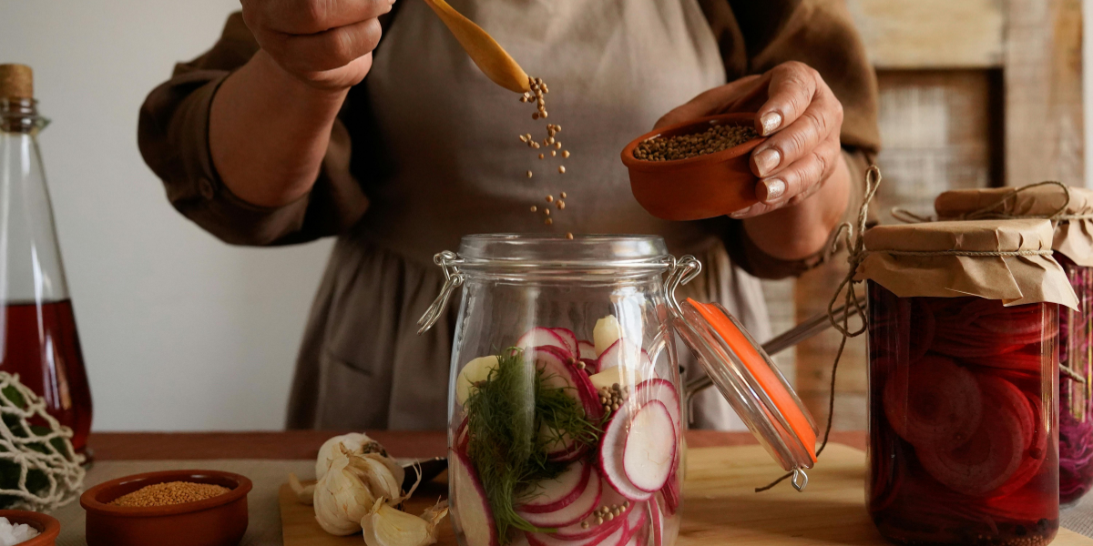 Person adding spices to a jar of pickled vegetables in a kitchen setting