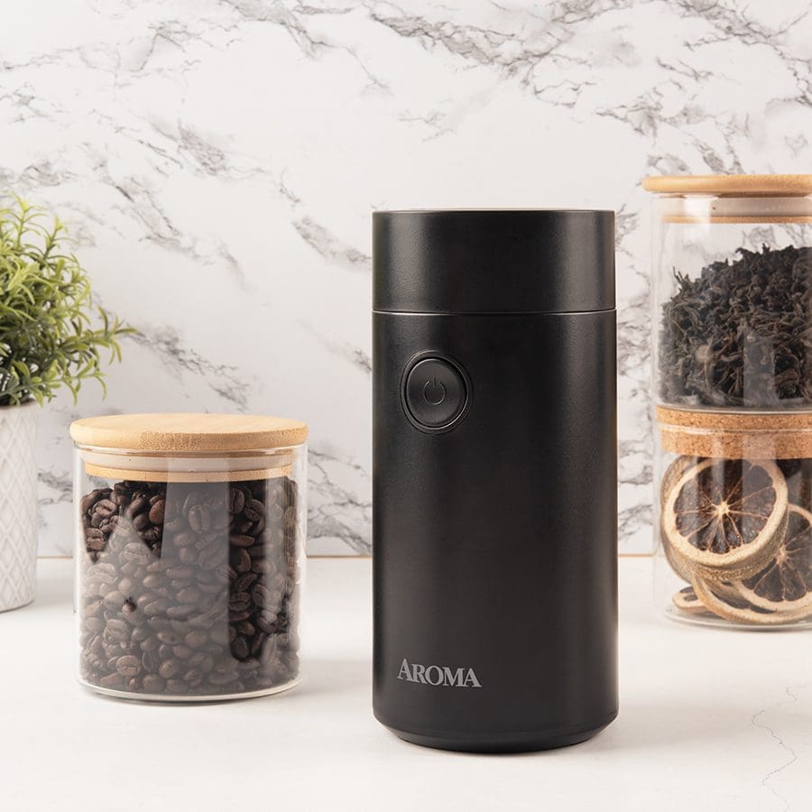 Black coffee grinder with labeled 'AROMA' on a marble surface with glass jars containing coffee beans and dried herbs.