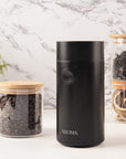 Black coffee grinder with labeled 'AROMA' on a marble surface with glass jars containing coffee beans and dried herbs.