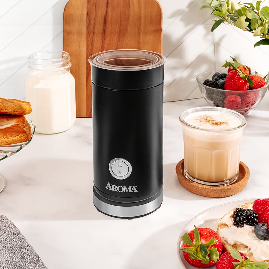Black Aroma milk frother on a kitchen counter with a glass of coffee and fruit bowl in the background.