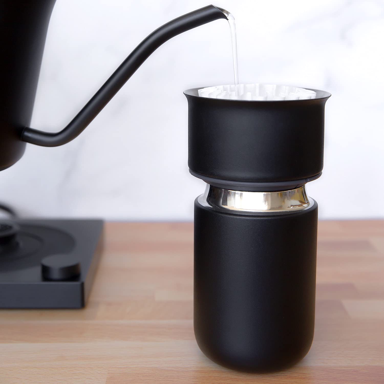 Black gooseneck kettle pouring liquid into a black travel mug on a wooden surface.