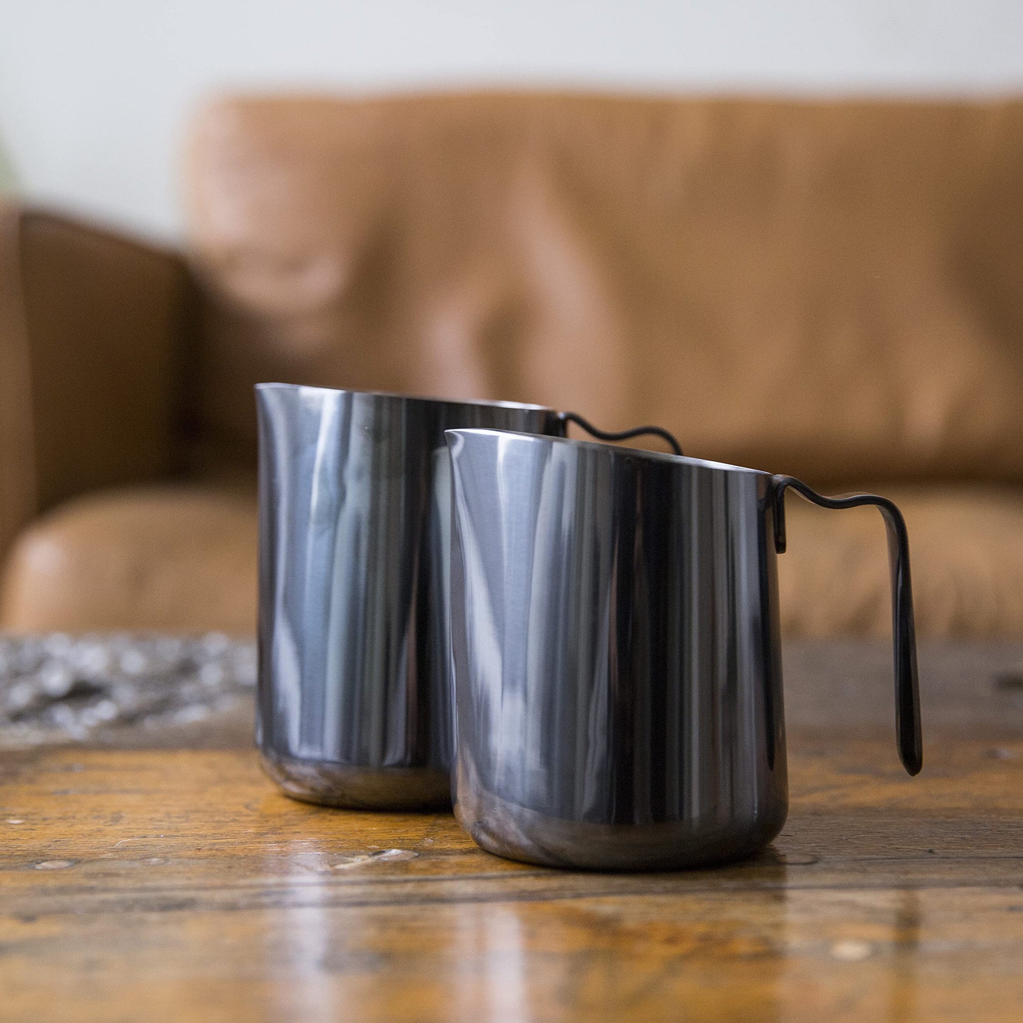 Two metallic milk steaming pitchers on a wooden table with a blurred brown couch in the background.