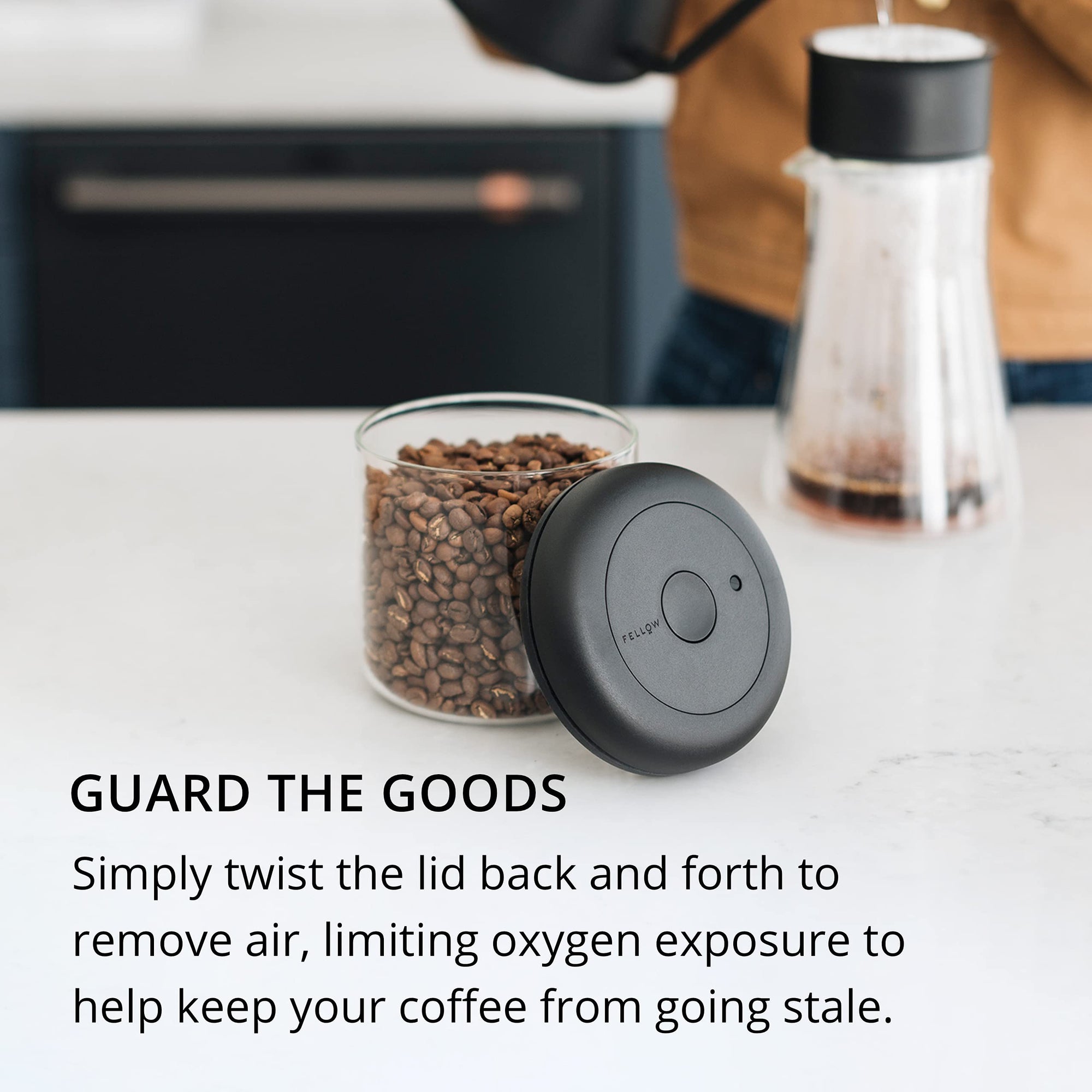 Glass container with coffee beans and a black lid on a kitchen counter with text about glass container.