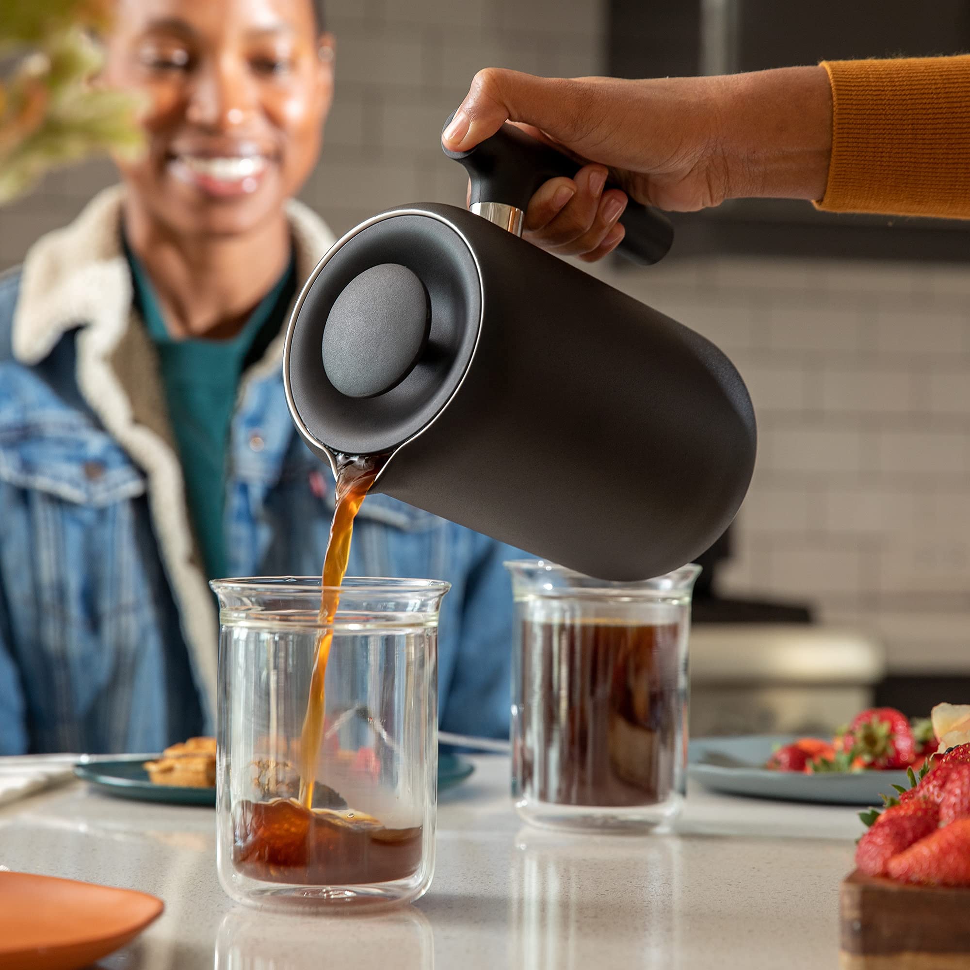 Person pouring coffee from a black french press into a glass, with another person in the background.