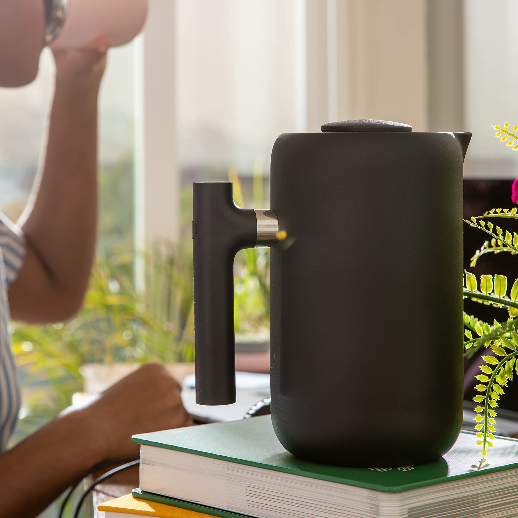 Black french press on a stack of books with a blurred person and plants in the background
