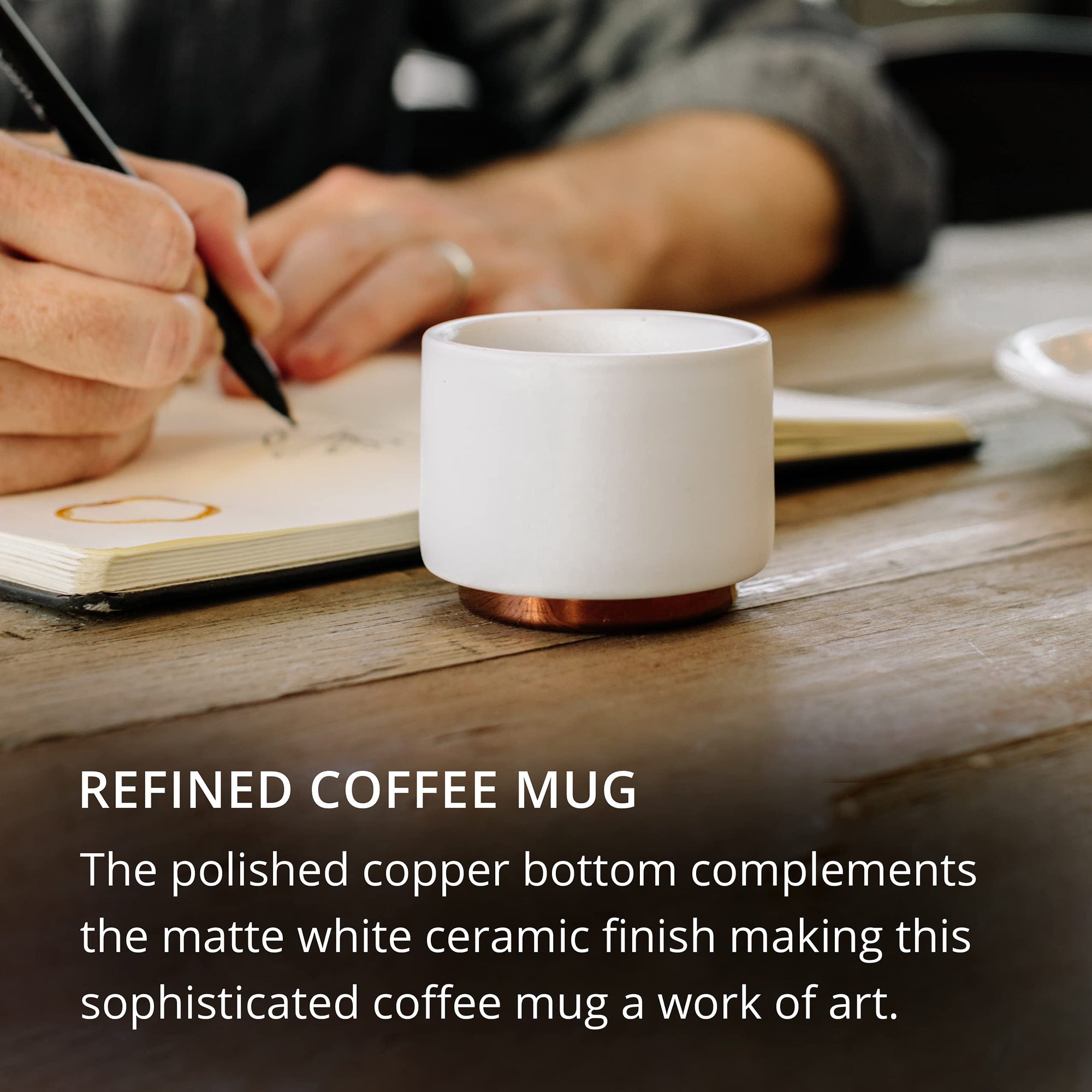 White coffee mug with copper bottom on a wooden table with hands writing in a notebook with text describing coffee mug.