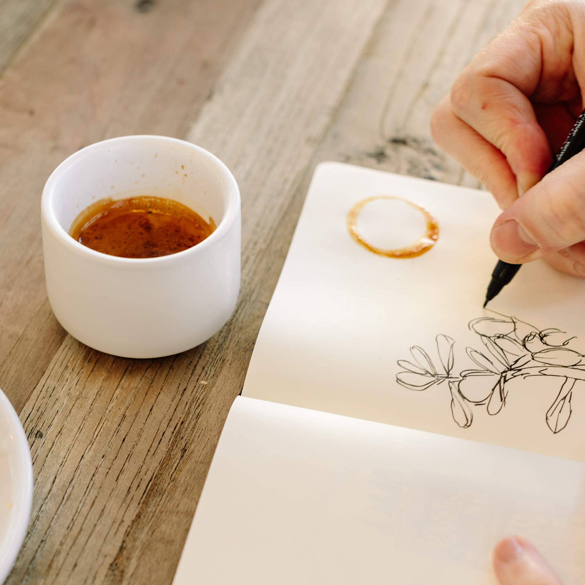 Person drawing on a piece of paper next to a small cup of coffee on a wooden table