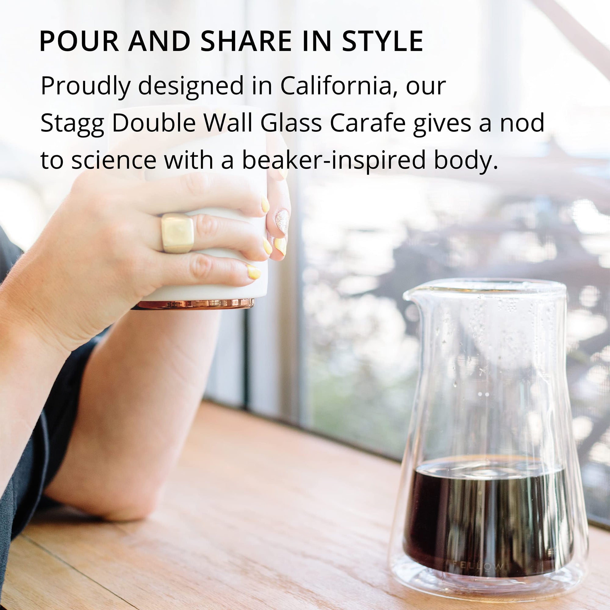 Stagg Double Wall Glass Carafe containing dark liquid inside with a person holding a cup of coffee indoors.