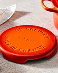 Flame Le Creuset enameled cast iron signature trivet on a wooden background with a kitchen towel and stockpot