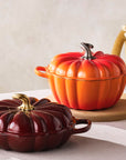 Two pumpkin-shaped cast iron cookware items on a table with pumpkins and a vase in the background.