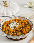 Baked dish with sweet potatoes and chickpeas on a white table with a cast iron pumpkin braiser and glasses.