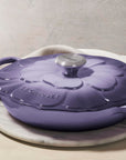 Purple cast iron cookware with floral design on lid on a marble surface with a white napkin and purple tulips.