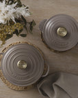 Two gray Le Creuset pots with gold knobs on a wooden surface with decorative elements.