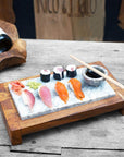A sushi server featuring a rectangular teak wood base with an inset marble top, presented with sushi rolls, sliced fish, and chopsticks on the marble surface.