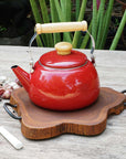 Rico & Plato amla teak trivet with a red kettle on top next to a spatula, shallots, garlic, and peppercorns on a wooden surface outside