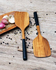 2 Rico & Plato bistro teak spatula with black handles on a wooden plate with shallots, garlic, herbs, and peppercorns on a wooden surface