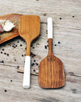 2 Rico & Plato bistro teak spatula with white handles next to a large wooden plate with herbs, garlic, shallots, and peppercorns on a wooden surface