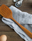 Rico & Plato emanuel teak spatula with air holes next to measuring spoons, bowl of salt, eggs, and laying on table cloth on a wooden surface