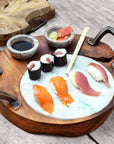 A teak wood sushi server with integrated handles and two gray marble pots, displayed with sushi and dipping sauce on a wooden surface.