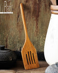 A wooden spatula with drainage slots, resting against a rustic background, part of the kitchen utensils collection by Rico & Plato.