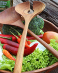 Two teak salad serving utensils placed on a wooden bowl with fruit and veggies on a wooden background