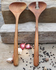 Two handcrafted teak wood salad servers with smooth, ergonomic handles, placed on a wooden surface surrounded by scattered black peppercorns and garlic cloves.