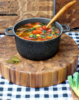 Rico & Plato Bayleaf and teak chopping board with a pot full soup and wooden spoon on a black and white checkered surface and end grain cutting boards in the back