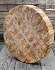 A round, teak wood end grain cutting board with a checkered pattern, displayed on a wooden surface.