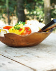A handcrafted teak wood salad bowl with a natural edge, placed on a wooden surface, filled with salad.