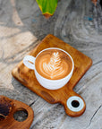 A rectangular teak wood coffee cup server with a white coffee cup placed on top, featuring a latte art design, served on a wooden surface.