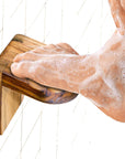 A person's foot with foam on it resting on a solid teak wooden shower footrest, placed on a white tiled wall.
