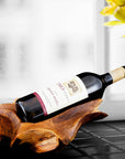 A teak wine bottle holder with an irregular shape, holding a wine bottle, placed on a table with a window in the background.