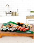 Rico & Plato wasabi teak serving platter with an assortment of sushi on top displayed on a light colored granite countertop in a kitchen setting