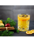 Riedel fine crystal tumbler spey whisky with a cinnamon and orange drink inside next to sliced oranges, cinnamon sticks, a cherry, and pine tree branches on a black surface