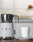 White Smeg retro line tritan renew milk frother next to a cup of milk on a wooden countertop