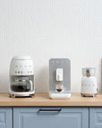 White Smeg retro line drip coffee maker, espresso maker, and milk frother on a wooden kitchen countertop next to milk with a white tile background
