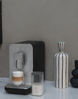 Black, silver Smeg fully automatic coffee machine with auto froth on a marble counter next to food, vases, and creamy coffee