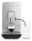 Black, silver Smeg fully automatic coffee machine with auto milk froth to the side and with a creamy espresso shot on a white background