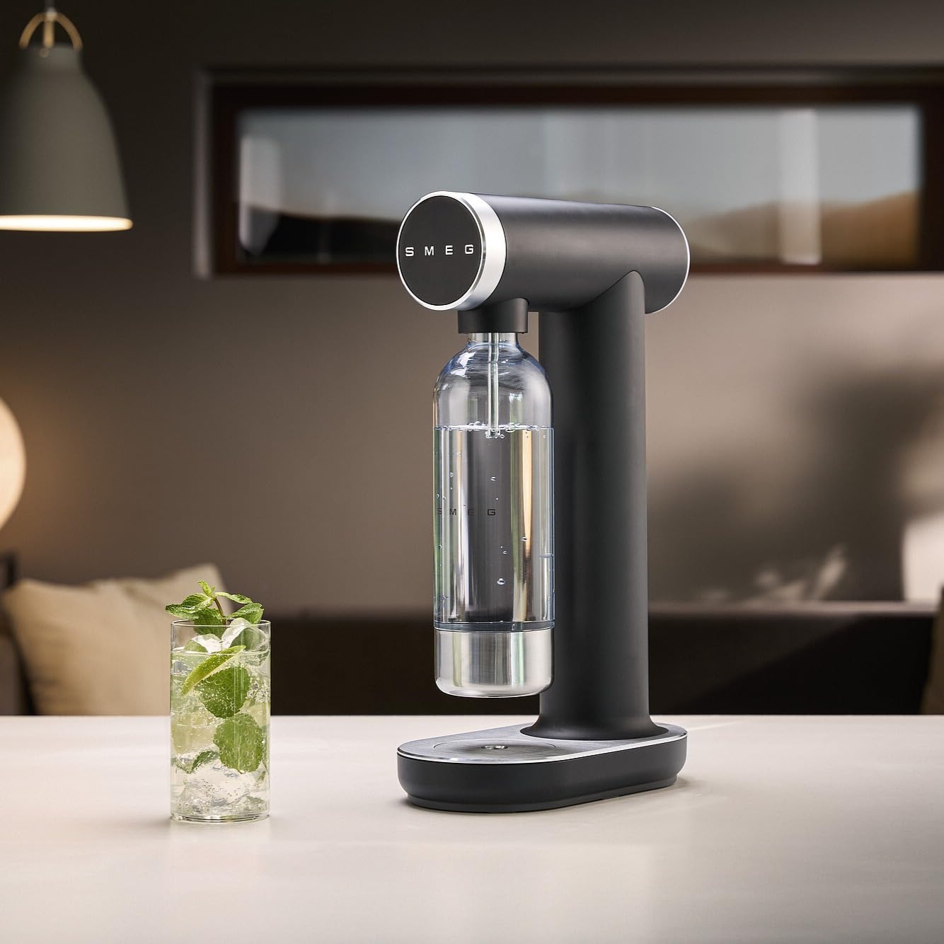 Black Smeg soda maker on a table with a glass of soda and mint leaves in a modern interior setting.