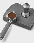 Grey, stainless steel Smeg tamper and portafilter with ground coffee grinds inside on a white background