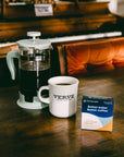 French press with coffee, a cup, and Third Wave Water Classic Light Roast Profile box on a wooden table in front of a piano