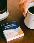 Hand holding a white mug with coffee, next to a box of 'Third Wave Water' and French press on a wooden table.