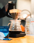 Coffee brewing setup with a person holding a coffee pot, a filter, and a box of Third Wave Water Dark Roast Profile on a wooden surface.