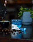 Box of Third Wave Water with coffee beans and a kettle pouring into a cup of coffee on a table.