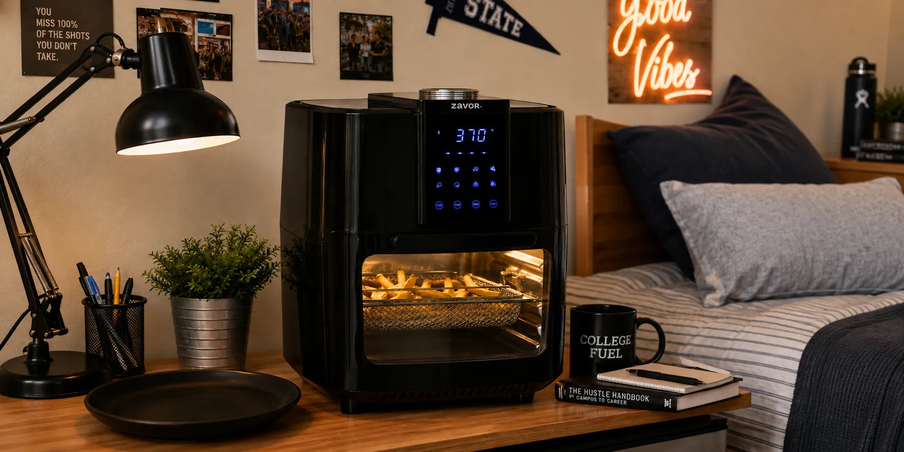 A black air fryer with a digital display window showing food cooking inside, placed on a wooden nightstand in a styled bedroom setting with a lamp, plant, bed, and decorative wall signs.