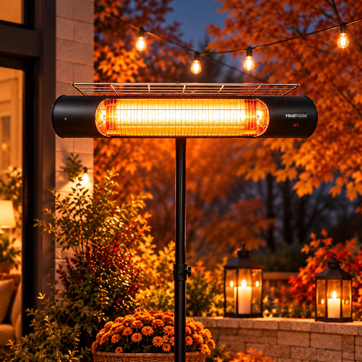 A black outdoor patio heater with a glowing orange infrared tube and a small metal shelf on top, shown standing on a post in a garden at dusk. The brand name 'HeatMate' is visible on the right side of the heater housing.