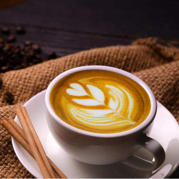 White cup and saucer with latte art and cinnamon sticks surrounded by coffee beans sitting on sacks and on a wooden surface