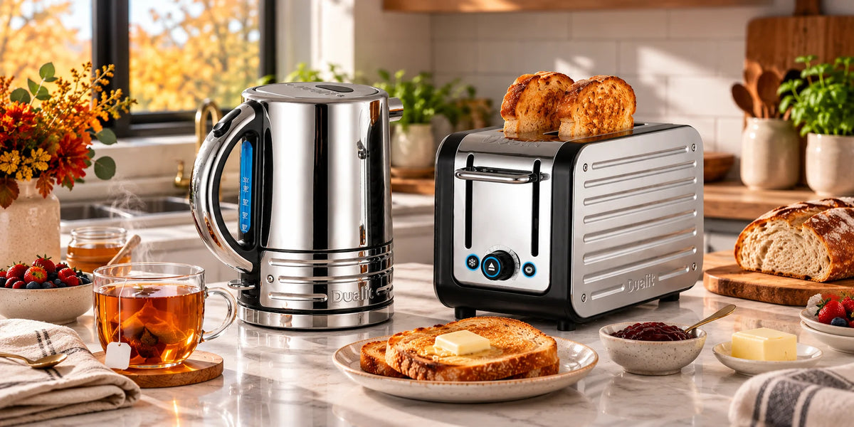 A stainless steel electric kettle and a silver-and-black toaster with two slices of toast, set on a kitchen counter surrounded by breakfast items, bread, fruit, and a warm autumnal window view.