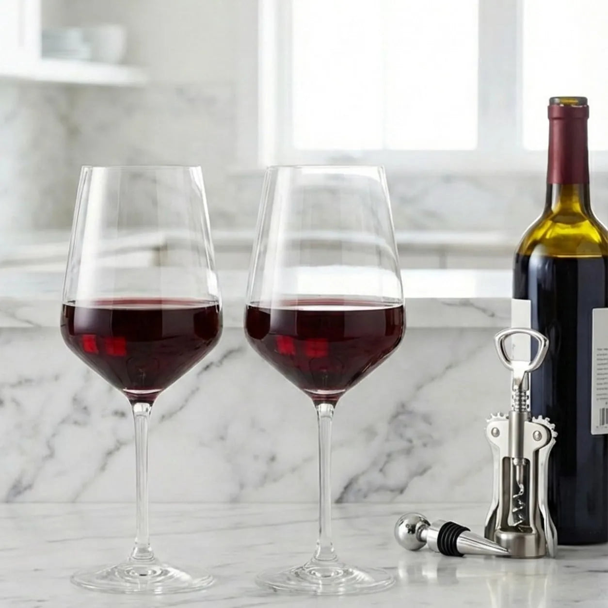 Two stemmed wine glasses filled with red wine on a white marble countertop, with a wine bottle and a silver corkscrew beside them in a bright kitchen setting.