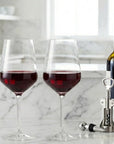 Two stemmed wine glasses filled with red wine on a white marble countertop, with a wine bottle and a silver corkscrew beside them in a bright kitchen setting.